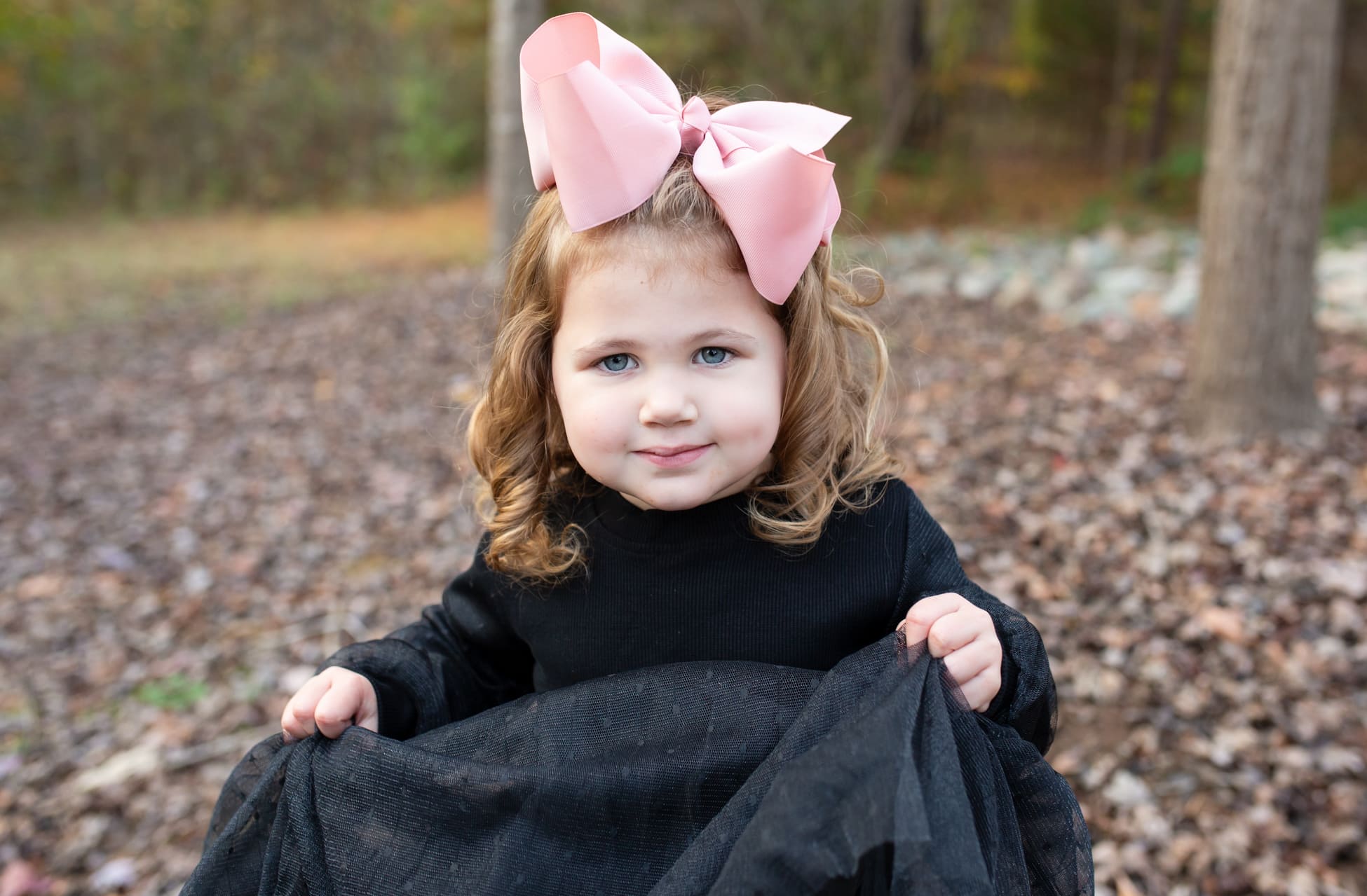a little girl wearing a black dress and cute pink bow on her head among fall leaves in Wake Forest, NC