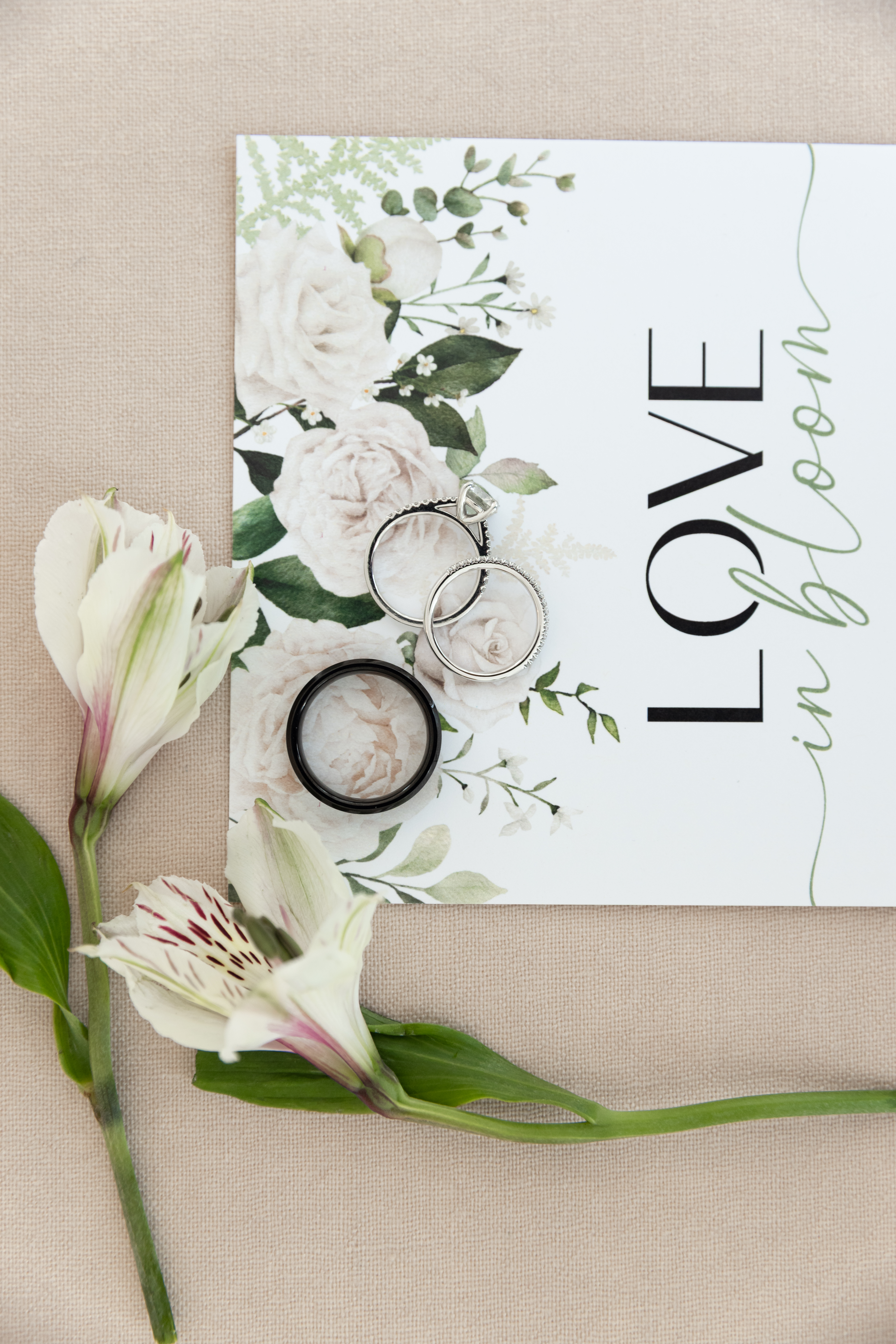 wedding bands on a wedding invitation featuring cloud dancer colored roses and greenery and words "love in bloom"