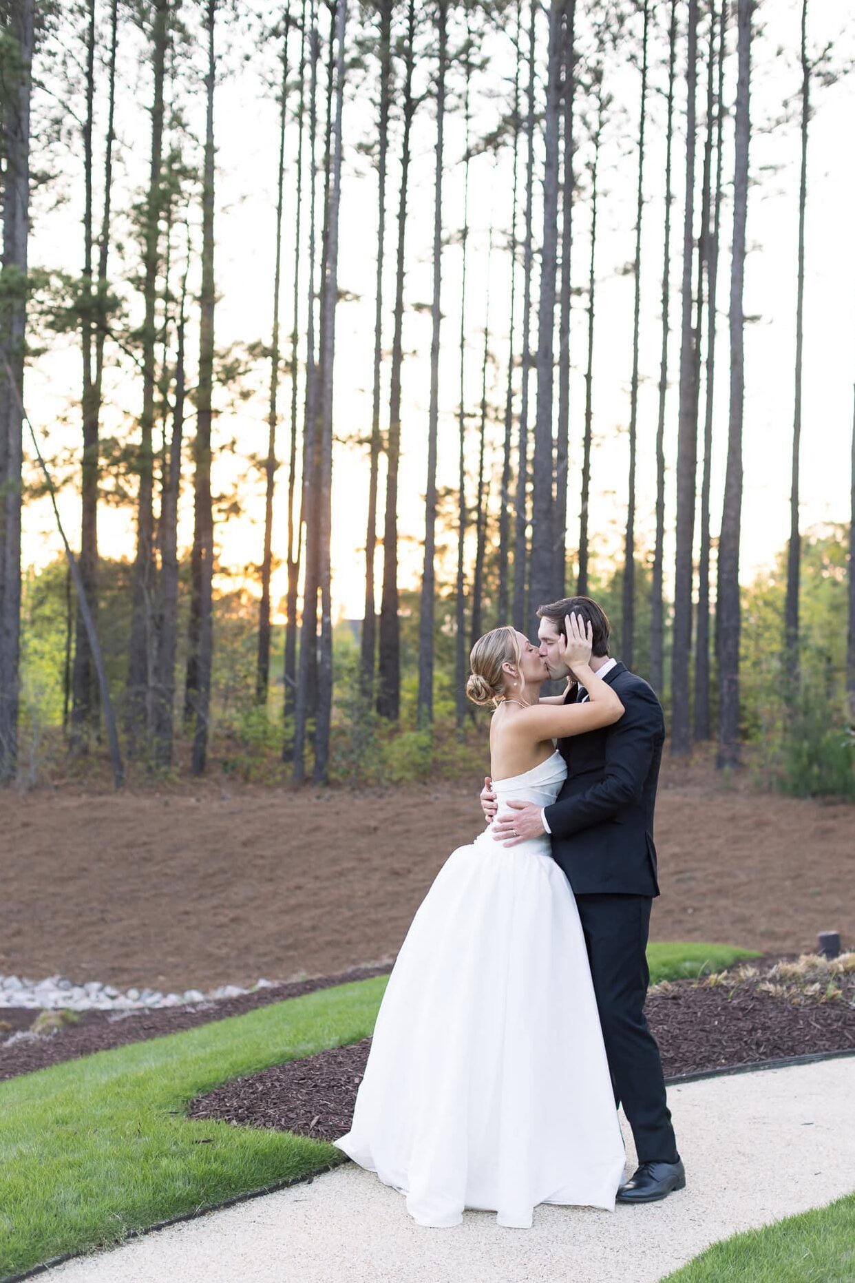 Newlyweds kissing, sun is setting behind tall pine trees in the background