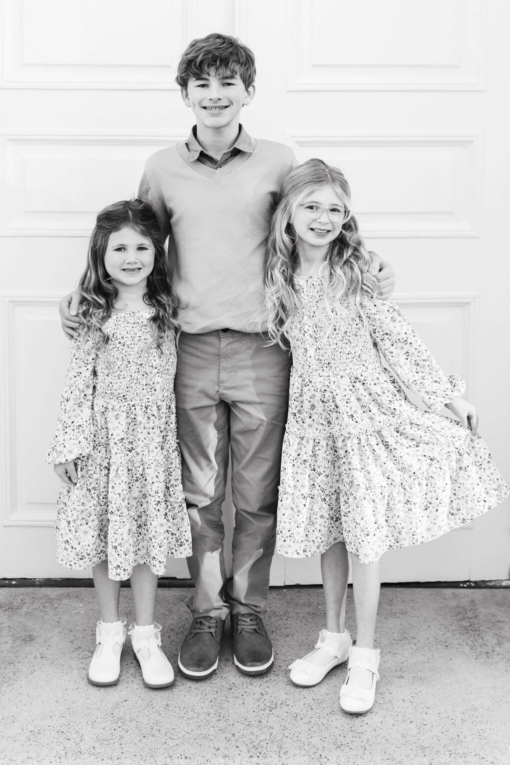 Black and white capture of 3 siblings hugging and posing for their christmas photo. the brother is in the middle and has his hands around the sisters shoulders. One sister is holding her dress, all kids are smiling.