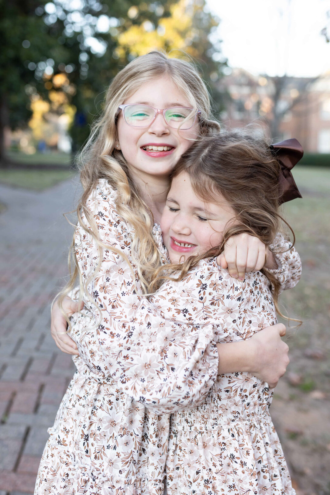 Two sisters wearing matching long sleeve, brown and beige floral dresses hugging and being silly during a fall mini session in Wake Forest. Wake Forest Mini Sessions: Relaxed Family Photos You'll Love for Years