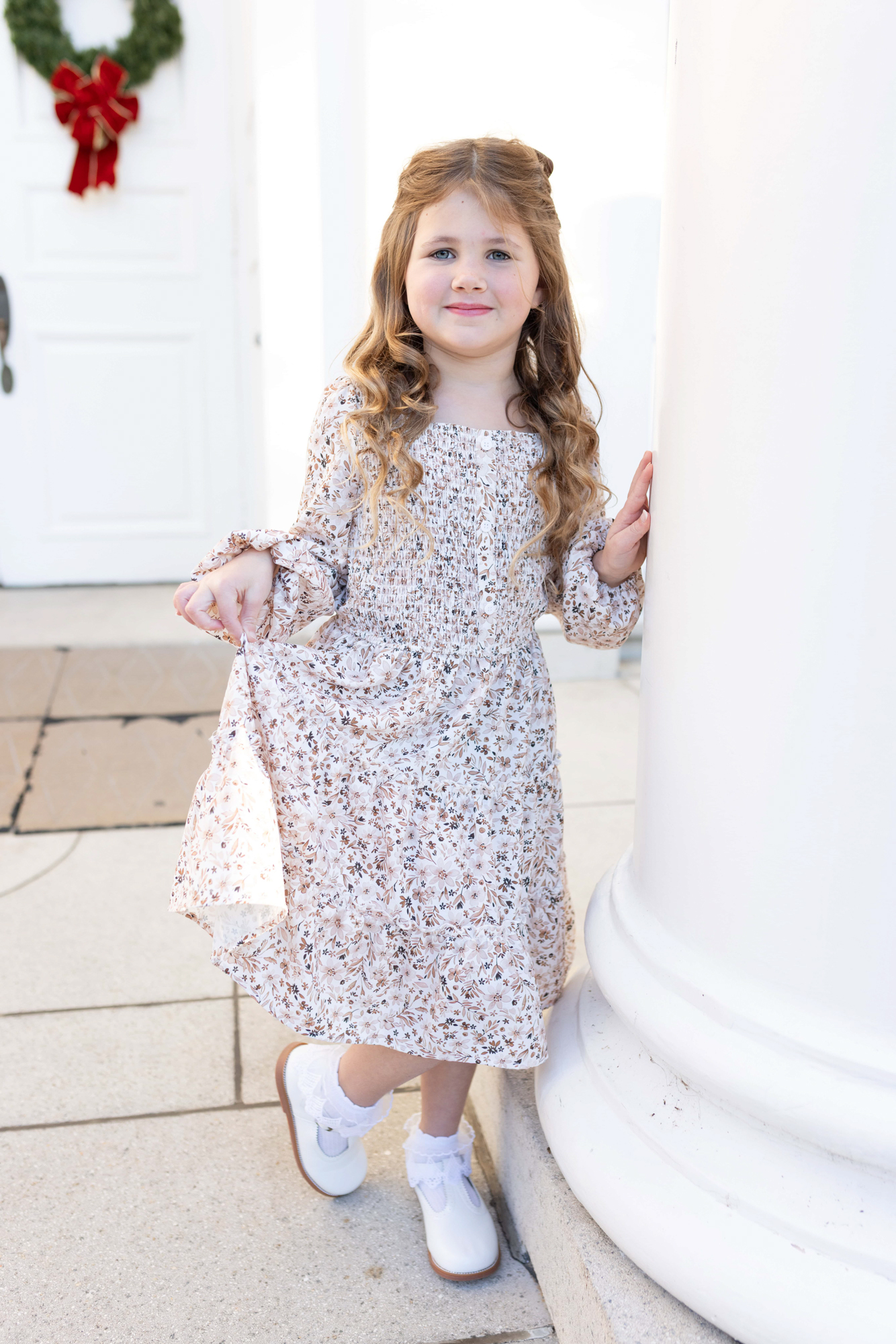 Little girl posing for a photo for a christmas card, in the background a white door with a christmas wreath hanging can be seen. Holiday photos Wake Forest, NC. Christmas mini sessions in wake forest, NC.